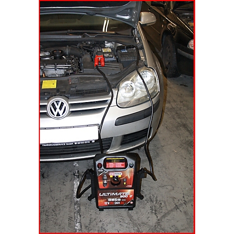 Ein silberner VW Golf mit geöffneter Motorhaube wird mit einem Starthilfegerät verbunden. Das Gerät steht vor dem Auto.